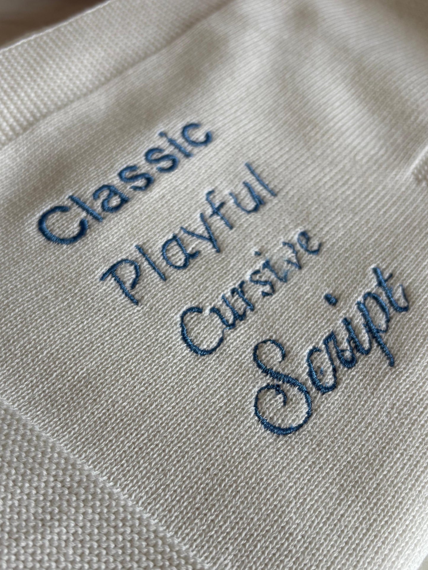 A close-up of a cream-colored woven fabric blanket with blue embroidered text reading 'Classic Playful Cursive Script.'