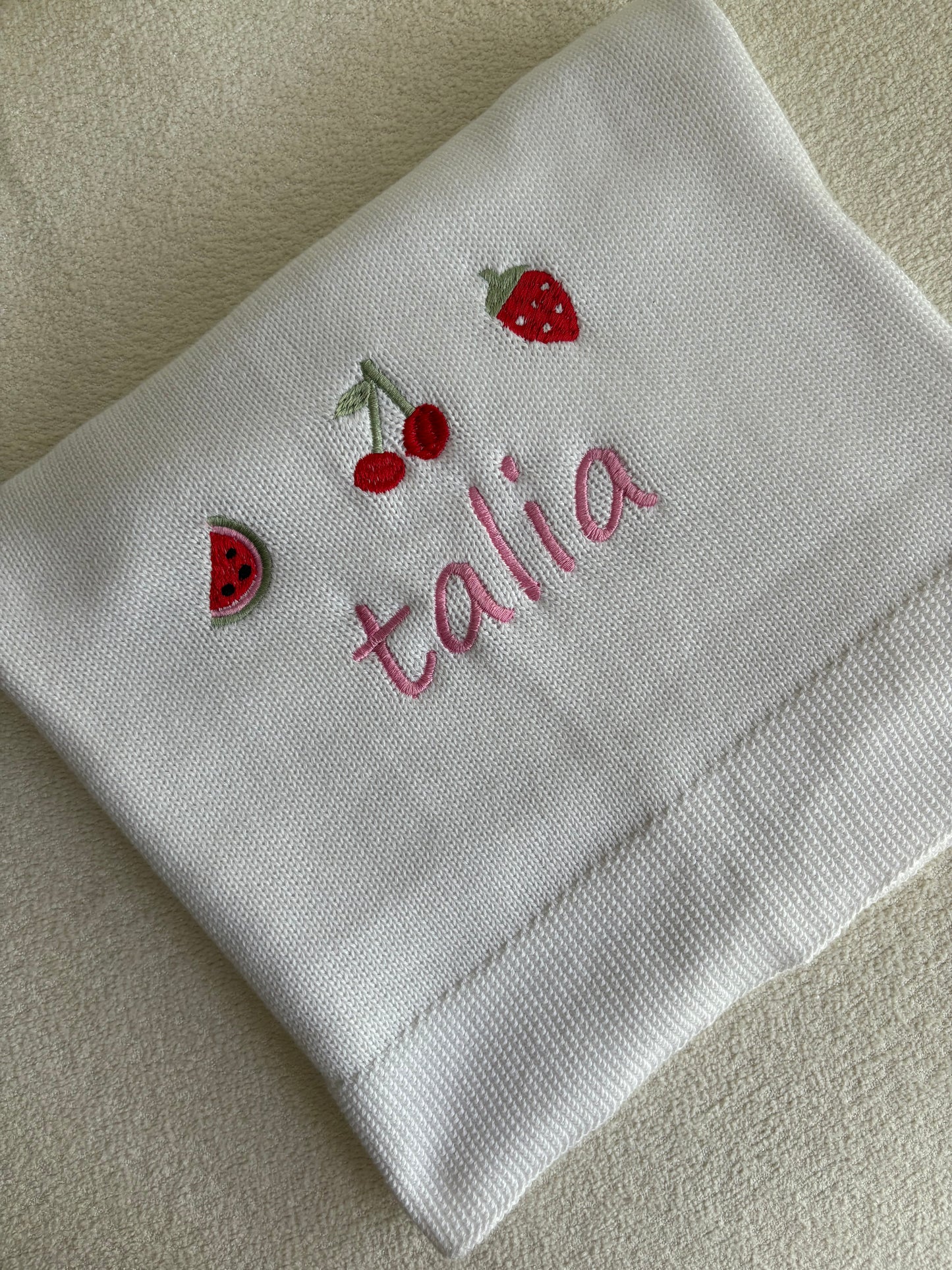 White folded blanket with embroidered name and strawberry designs on a beige background