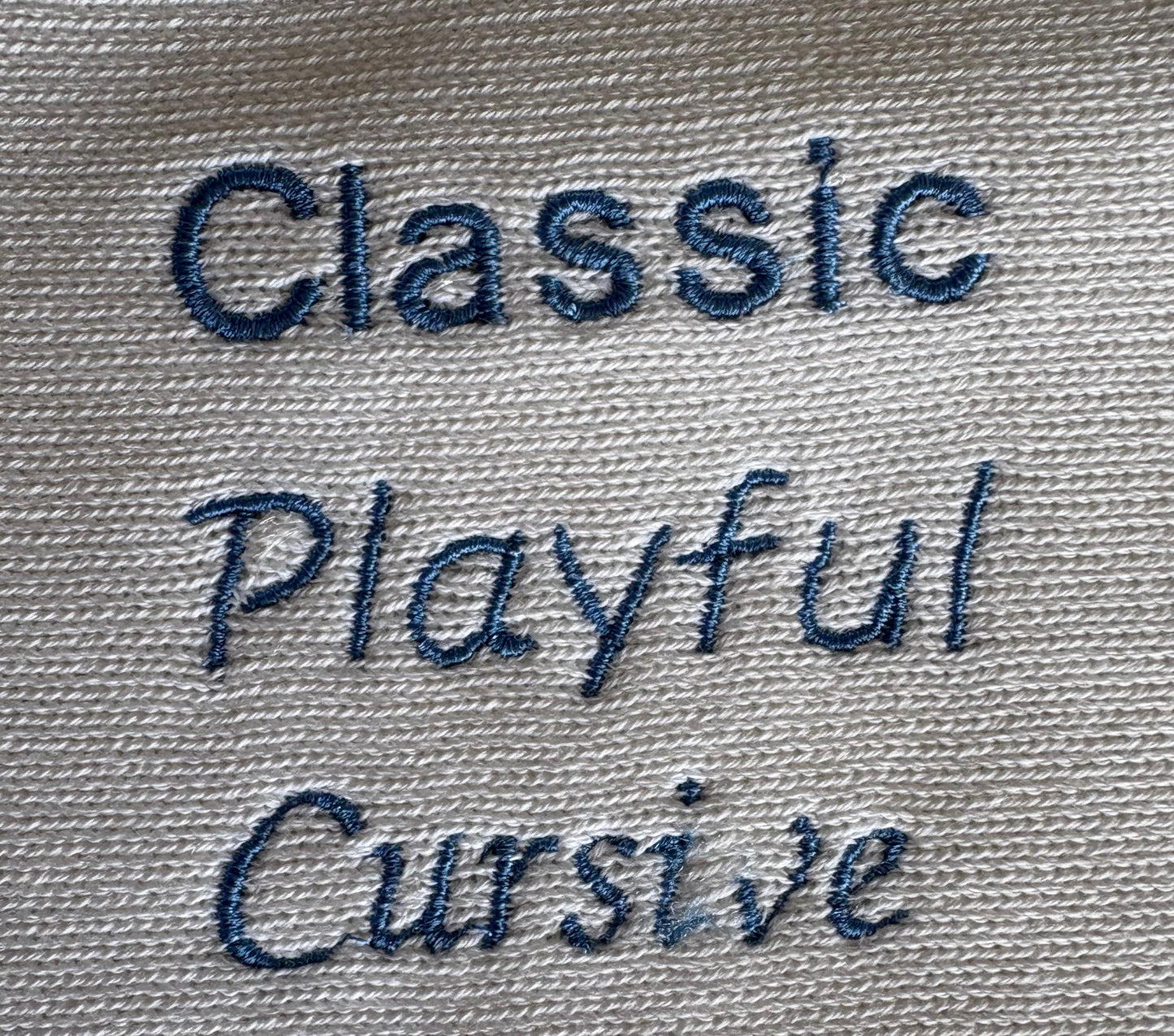 Close-up of embroidered text reading 'Classic Playful Cursive Script' in dark blue thread on a light white cotton knit blanket