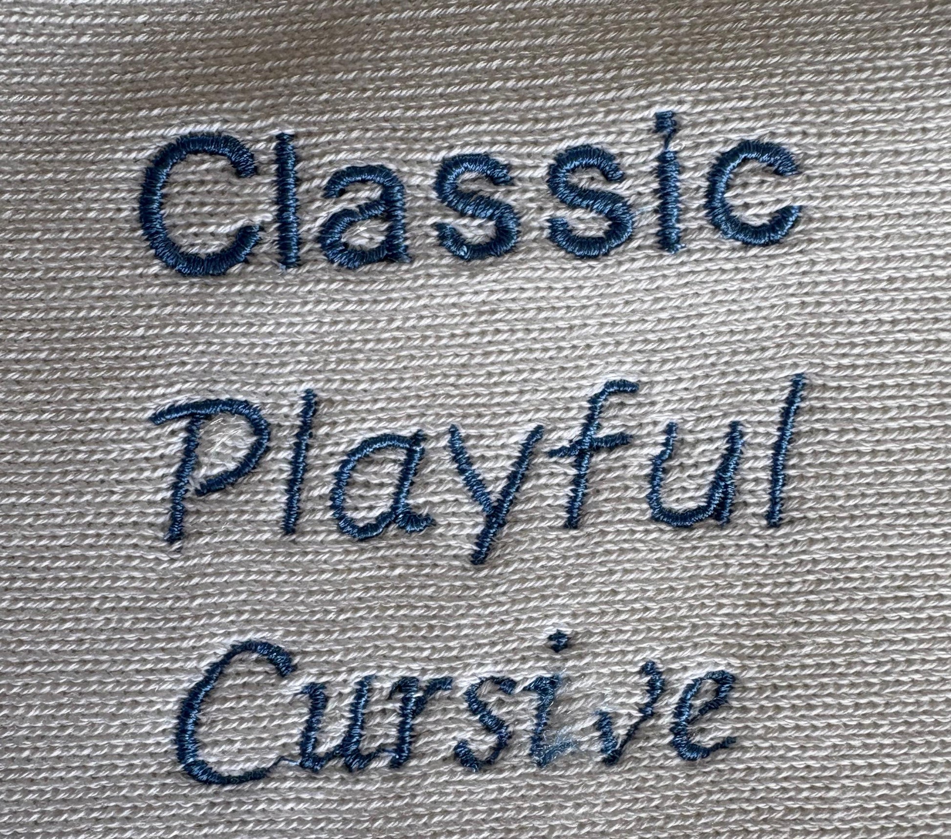 Close-up of embroidered text reading 'Classic Playful Cursive Script' in dark blue thread on a light white cotton knit blanket
