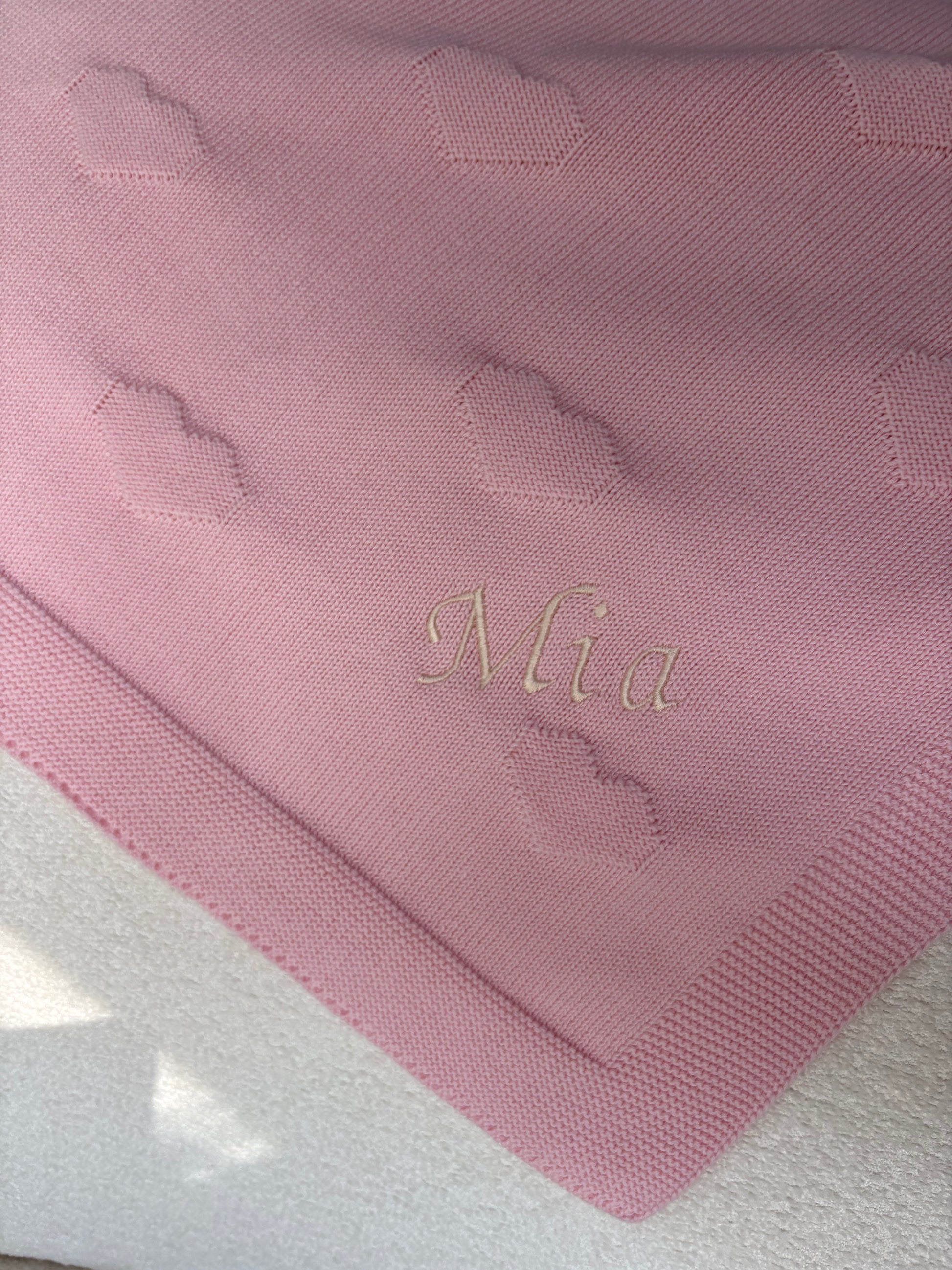 A close-up of a pink knit blanket featuring a textured heart pattern and a personalized name, 'Mia', embroidered in cream coloured thread in a cursive font.