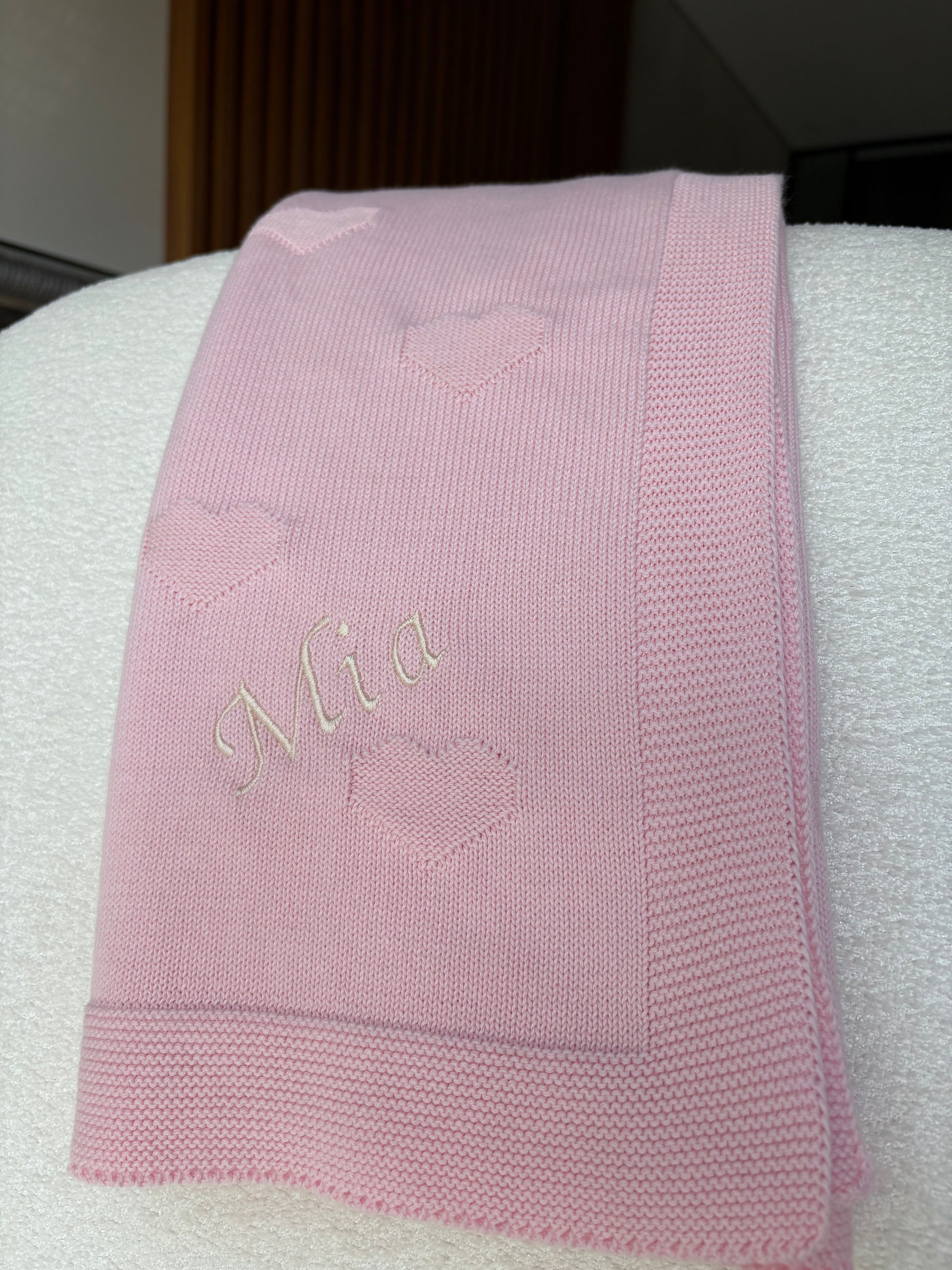 A folded light pink knit baby blanket with embroidered hearts and the name 'Mia' in cream cursive script font, displayed on a white textured surface.