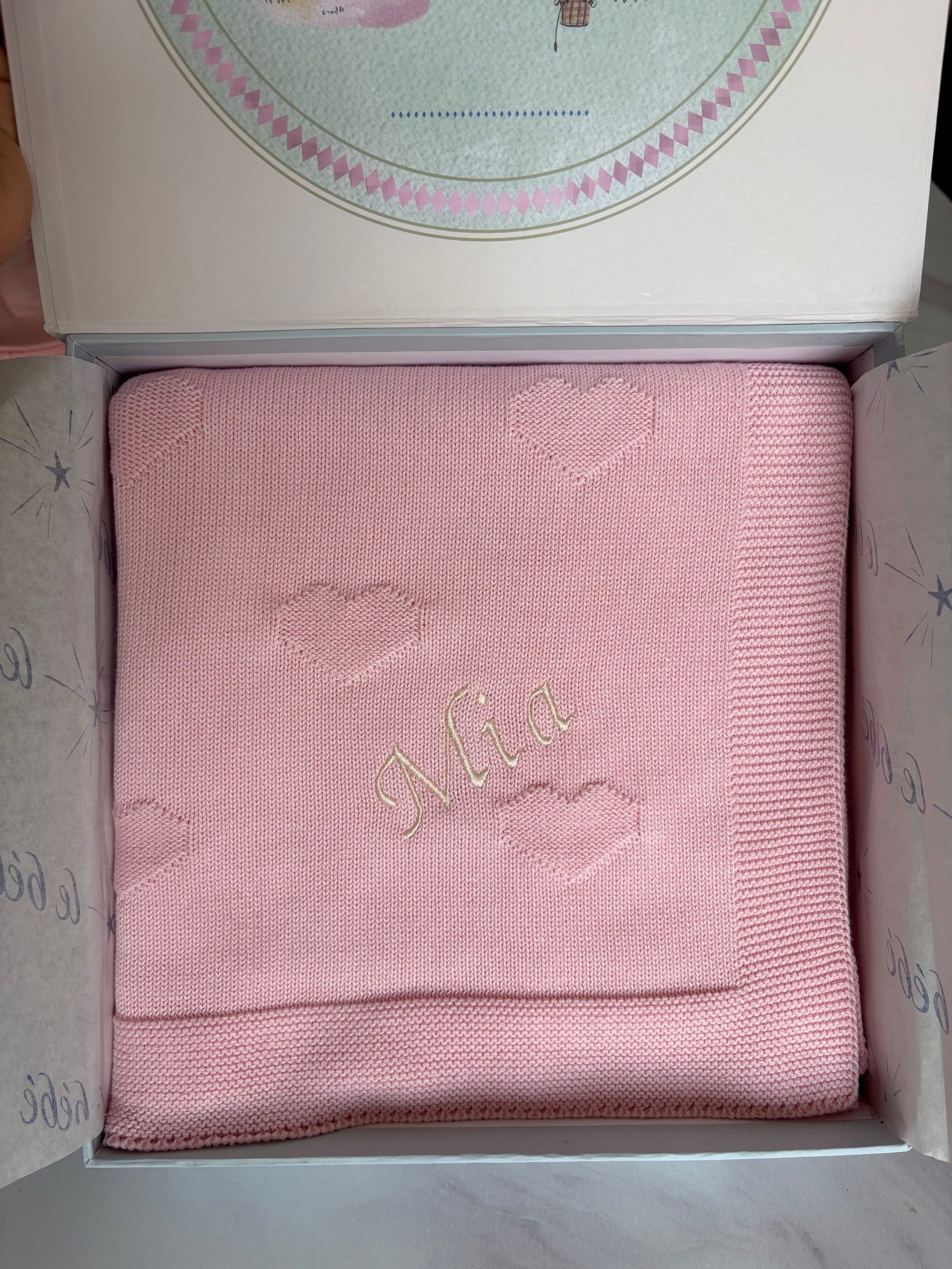 A folded light pink knitted baby blanket with embroidered hearts and the name 'Mia' in cream, presented inside a special keepsake gift box.