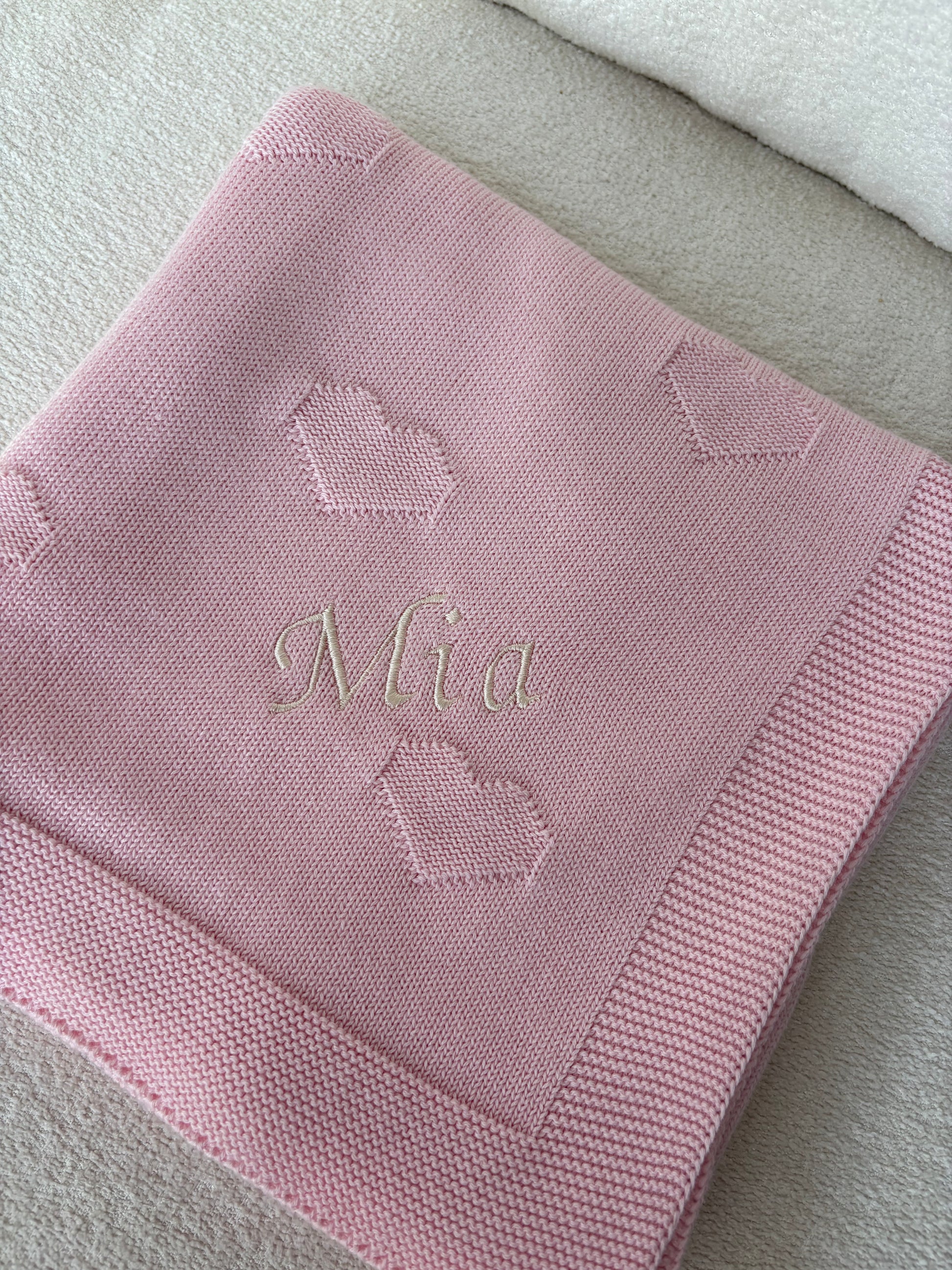 A pink knitted baby blanket with a stitched border, embroidered heart motifs, and the name 'Mia' in cursive font, laid folded on a neutral cream surface.