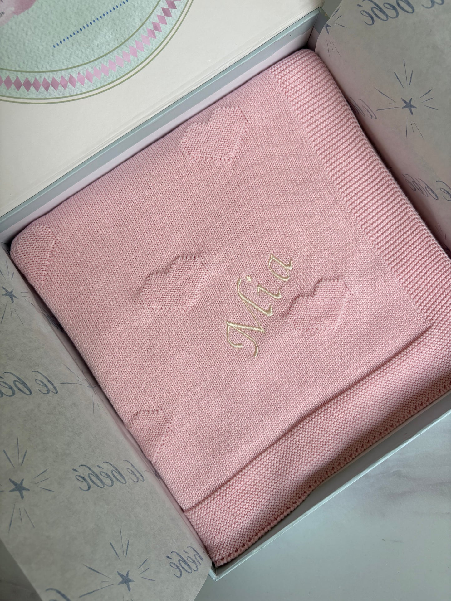 A folded light pink baby blanket with embroidered cloud motifs and the name 'Mia' in cream cursive font, presented inside a gift box with le bebe patterned tissue paper.
