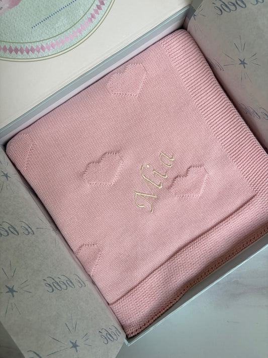 A folded light pink baby blanket with embroidered cloud motifs and the name 'Mia' in cream cursive font, presented inside a gift box with le bebe patterned tissue paper.