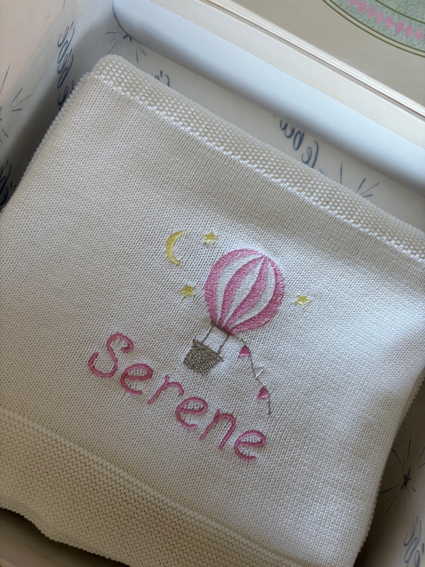 Embroidered white cotton knit blanket with a pink hot air balloon motif and personalised with the name 'Serene' in pink thread in a playful font. 