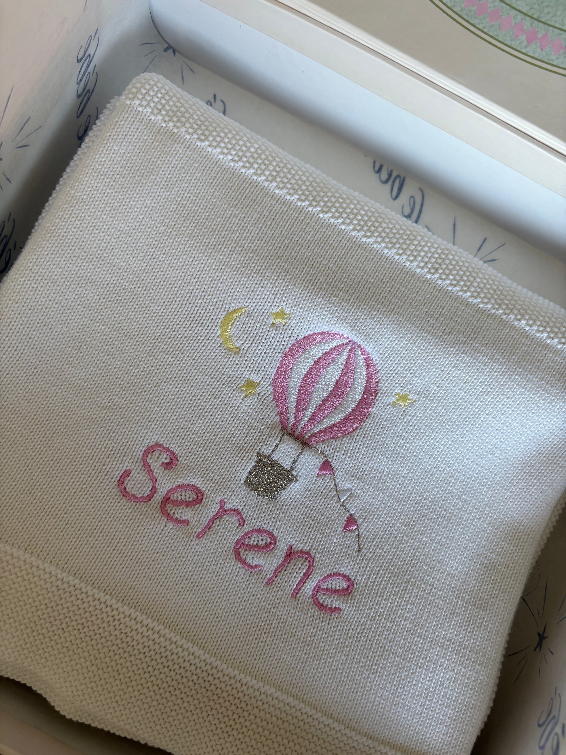 Embroidered white cotton knit blanket with a pink hot air balloon motif and personalised with the name 'Serene' in pink thread in a playful font. 