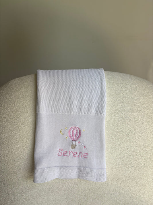White cotton knit blanket with embroidered design and 'Serene' in pink thread on a sofa. 